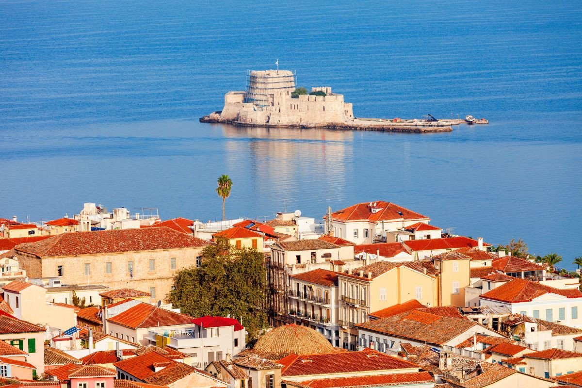 Colorful rooftops of Nafplio town with a historic fortress on a small island in the background, surrounded by calm blue waters.